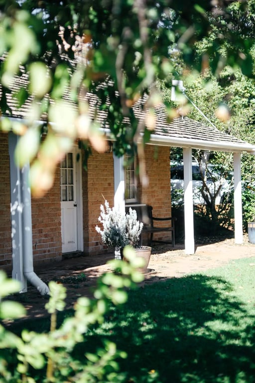 A charming brick cottage with a white verandah, nestled among green foliage, possibly on the Gingin Grass Fed property in WA.