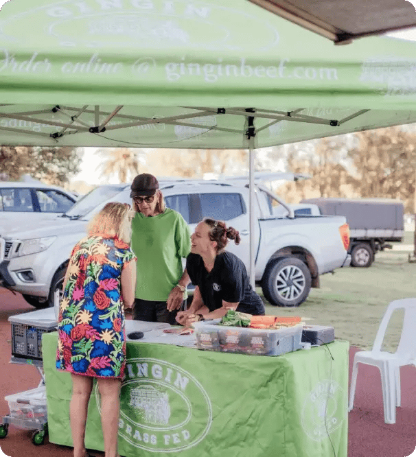 People interacting at the Gingin Grass Fed market stall in Gingin, WA, with branded signage visible.